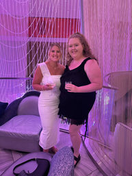 Two smiling women in a pink-lit lounge, one wearing a white sash and fitted dress and the other in a black ruffled dress, holding cocktails in front of beaded curtain decor.