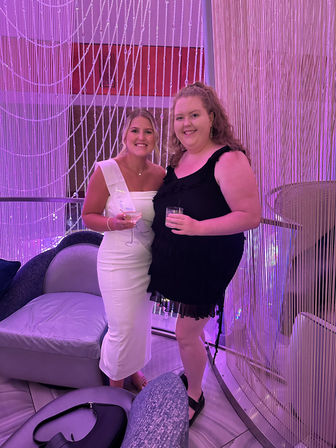 Two smiling women in a pink-lit lounge, one wearing a white sash and fitted dress and the other in a black ruffled dress, holding cocktails in front of beaded curtain decor.