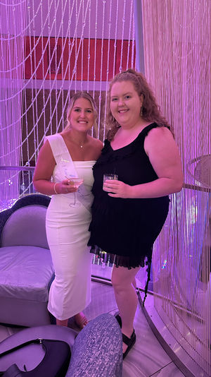Two smiling women in a pink-lit lounge, one wearing a white sash and fitted dress and the other in a black ruffled dress, holding cocktails in front of beaded curtain decor.