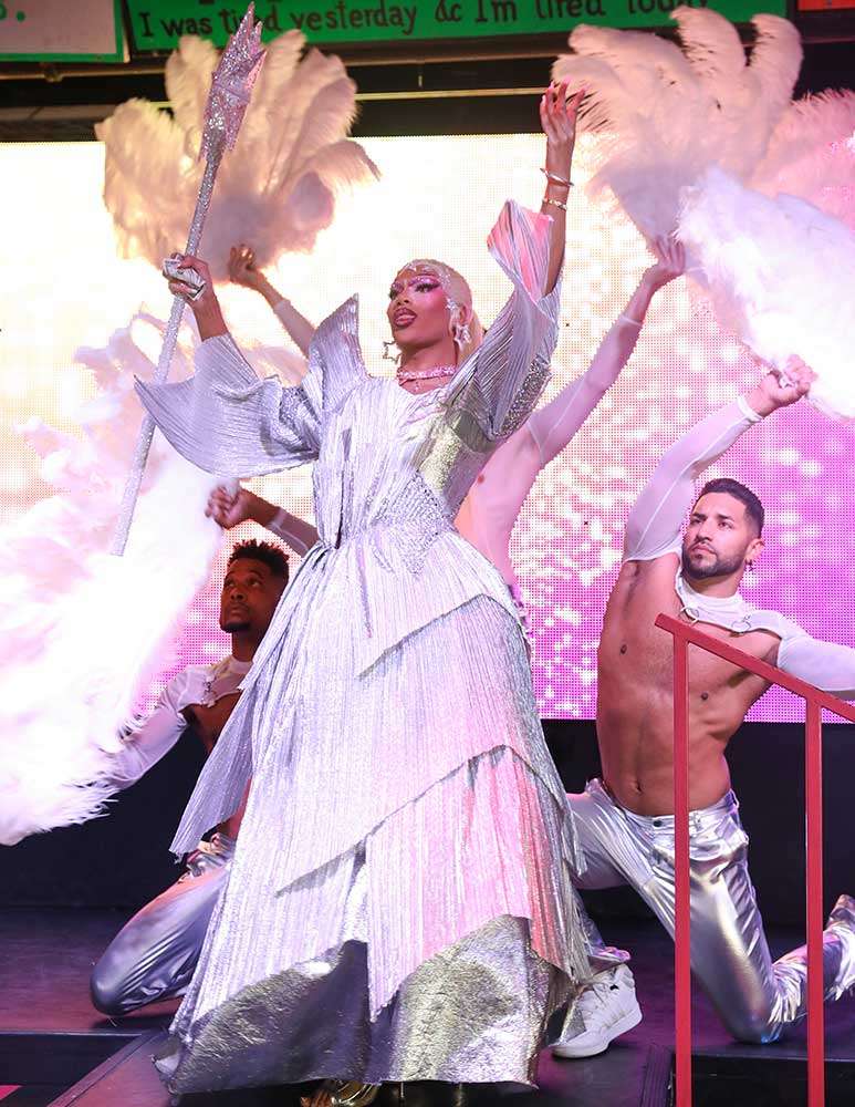 Drag performer in a shimmering silver gown holding a jeweled scepter on a cabaret stage, flanked by shirtless backup dancers waving large white feather fans against a glittering pink backdrop.