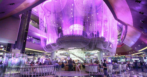 Vibrant Las Vegas casino interior featuring a multi-level central bar beneath a massive purple-lit beaded chandelier, surrounded by gaming floor and patrons.
