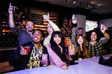 Group of friends cheering with cocktails and cans at a lively indoor bar, smiling at a white marble counter with an American flag and TVs in the background.