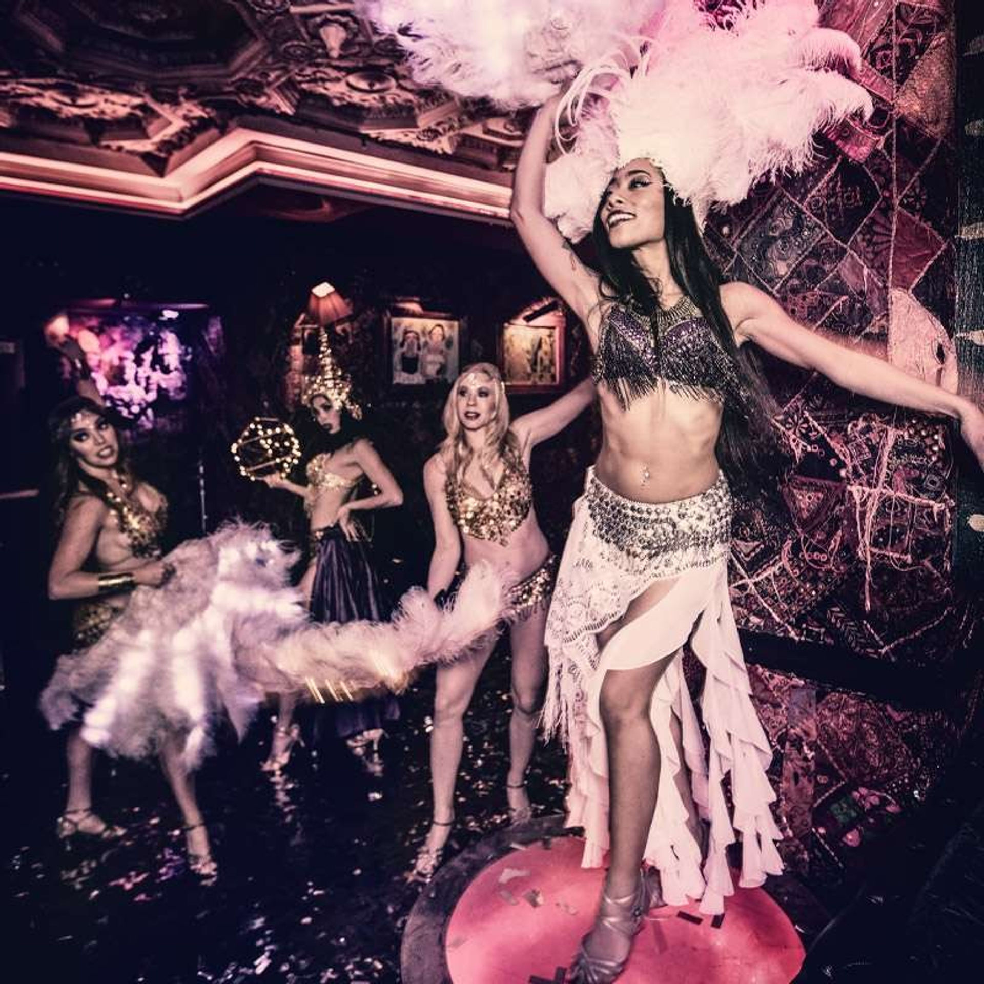 Glamorous burlesque dancers on a nightclub stage in sequined costumes and oversized feather headdresses, mid-performance.