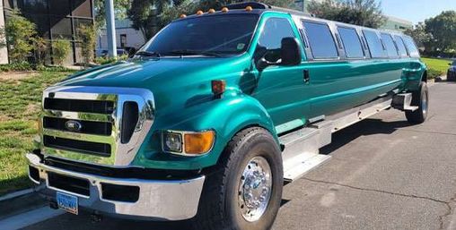 Turquoise-green stretched truck limousine with chrome grille and wheels and a long row of passenger windows, parked on a sunny suburban street.