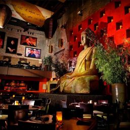 Candlelit Asian restaurant interior with a giant seated Buddha statue against a red textured wall, exposed brick, hanging lanterns, bamboo plants and cozy dining tables.