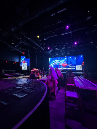 Indoor entertainment venue with a lit stage and large LED screen, purple and blue spotlights, audience seated at long tables, some wearing hats