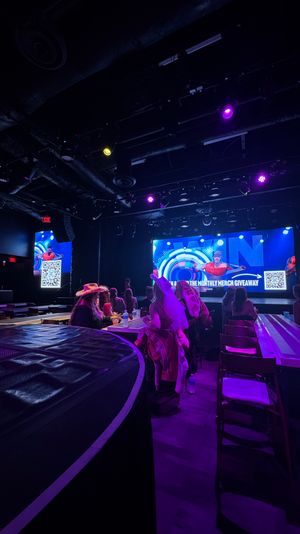 Indoor entertainment venue with a lit stage and large LED screen, purple and blue spotlights, audience seated at long tables, some wearing hats