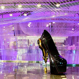 Glossy black high-heeled shoe sculpture with gold hardware beneath a cascading purple string-light chandelier in a modern hotel lobby with a reflective floor