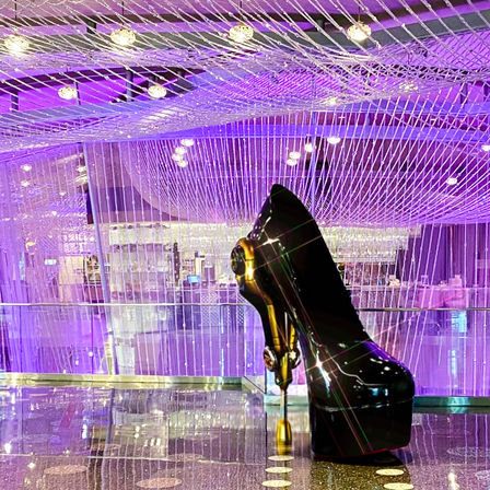 Glossy black high-heeled shoe sculpture with gold hardware beneath a cascading purple string-light chandelier in a modern hotel lobby with a reflective floor
