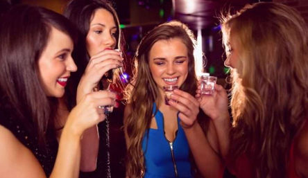 Four friends clinking shot glasses in a lively nightclub under colorful lights