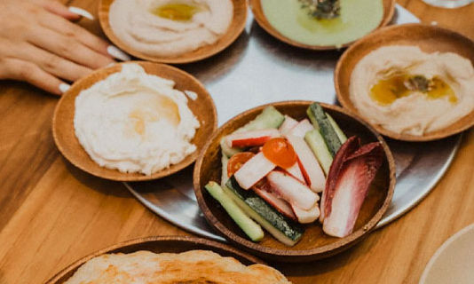 Mediterranean mezze on a wooden table: small wooden bowls of creamy hummus drizzled with olive oil, a bowl of cucumber, radicchio and cherry tomato crudités, and warm pita in wooden dishes.