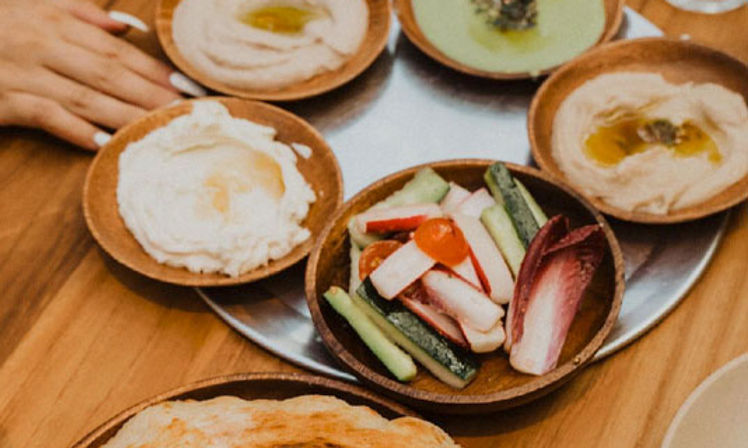 Mediterranean mezze on a wooden table: small wooden bowls of creamy hummus drizzled with olive oil, a bowl of cucumber, radicchio and cherry tomato crudités, and warm pita in wooden dishes.
