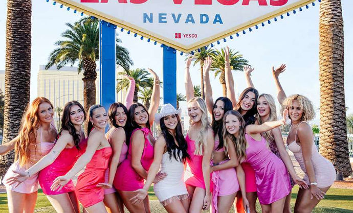 Group of women in pink and white party dresses posing and cheering in front of the Welcome to Fabulous Las Vegas, Nevada sign with palm trees in the background