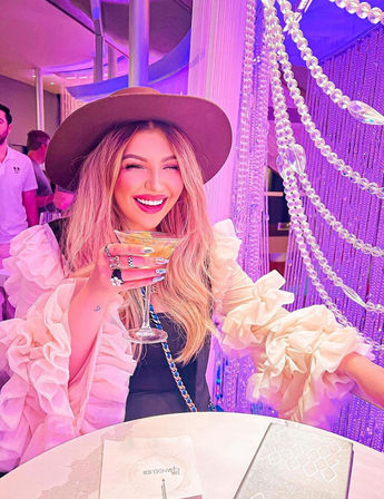 Smiling woman in a wide-brim hat and ruffled sleeves raising a coupe cocktail in a purple-lit, chandelier-adorned upscale lounge — vibrant nightlife scene