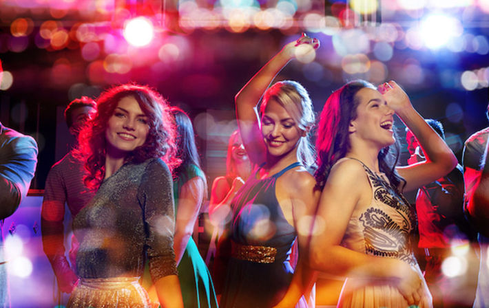 Three women dancing and laughing on a colorful nightclub dance floor under vibrant bokeh lights, lively city nightlife scene