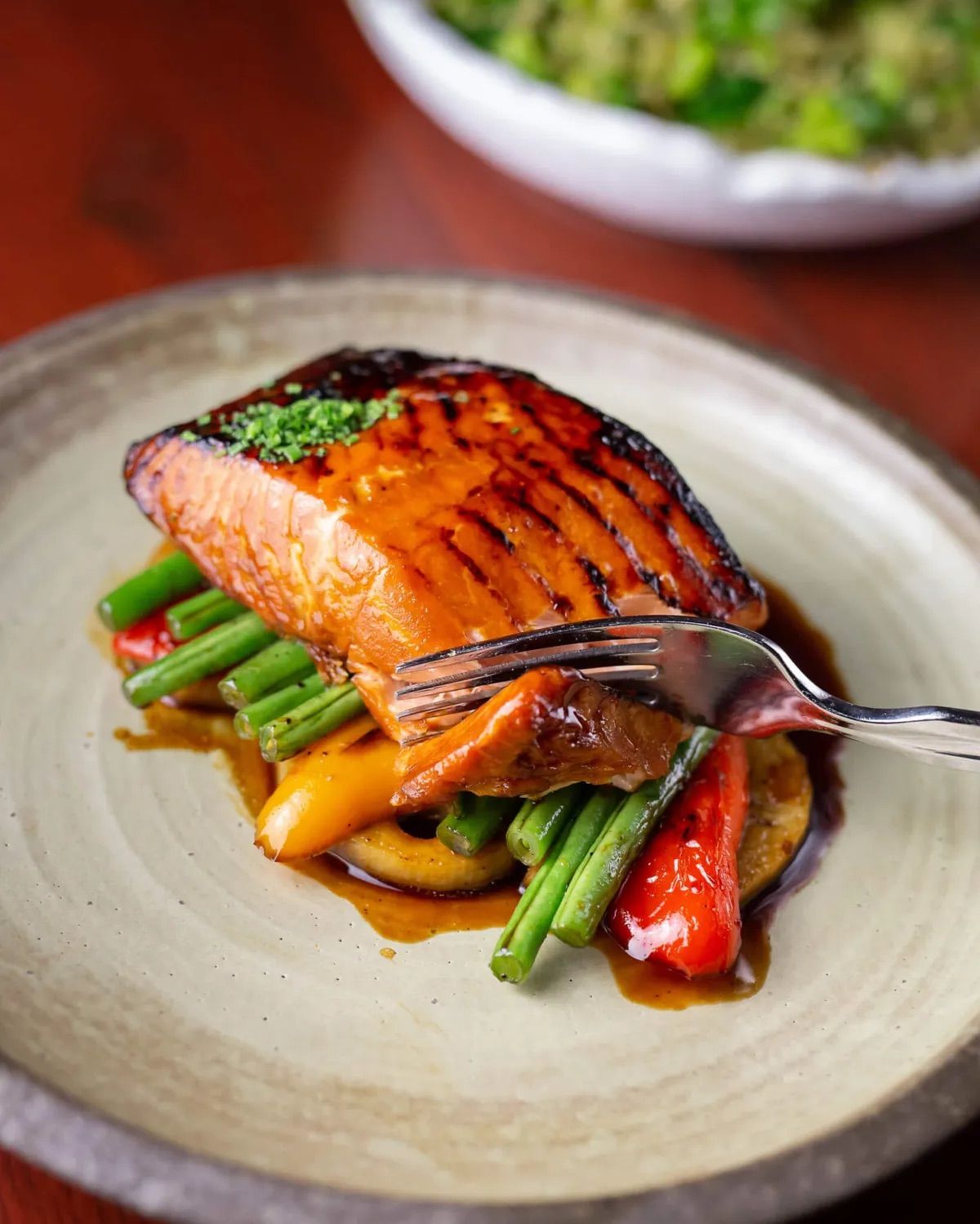 Caramelized grilled salmon fillet glazed in a brown sauce, served on green beans and roasted red and yellow peppers on a rustic ceramic plate with a fork flaking a bite