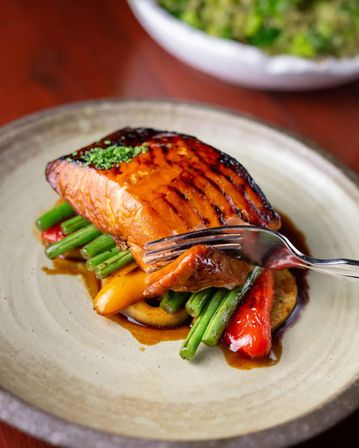 Caramelized grilled salmon fillet glazed in a brown sauce, served on green beans and roasted red and yellow peppers on a rustic ceramic plate with a fork flaking a bite