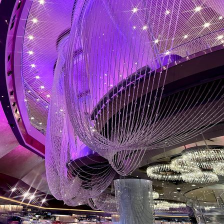 Giant draped crystal-bead chandeliers cascading from a high ceiling, bathed in purple mood lighting in a modern luxury lobby/atrium.