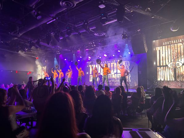 Shirtless male dancers in jeans tipping cowboy hats on a brightly lit cabaret stage while a seated audience films and cheers under purple spotlights