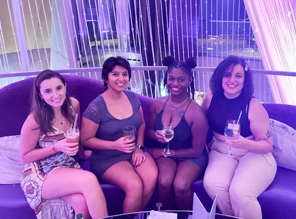Four women smiling on a curved purple booth in a purple-lit indoor lounge, holding cocktails and wine glasses in front of a beaded curtain — casual night out.