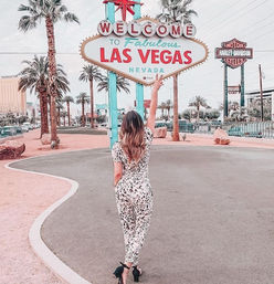 Rear view of a woman in a patterned jumpsuit raising her arm toward the iconic Welcome to Fabulous Las Vegas Nevada sign, framed by palm trees and a pastel-toned Strip backdrop.
