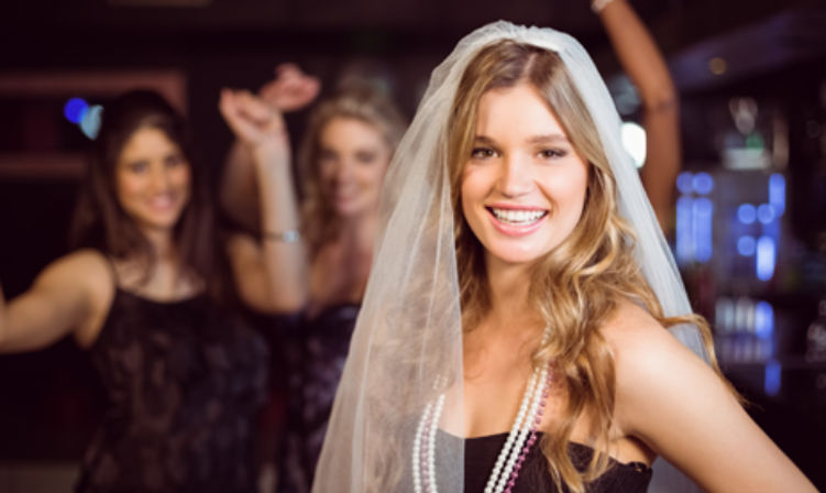 Smiling bride-to-be wearing a veil and layered pearl necklaces posing at a lively bachelorette party in a dimly lit bar with friends dancing in the background