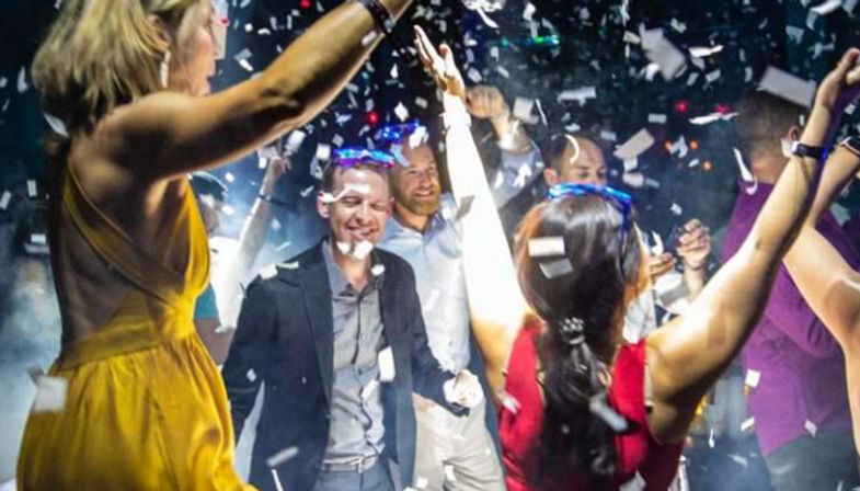 Group of people dancing and cheering under falling confetti on a lively nightclub dance floor with colorful lights