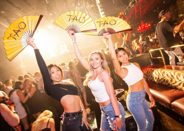 Nightclub party with three women in crop tops and jeans holding yellow folding fans aloft on a crowded dance floor under bright stage lights