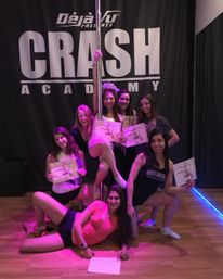 Group of women celebrating a pole fitness class graduation in a dance studio, posing around a vertical pole on a wooden floor under pink-purple lighting and holding certificates in front of a large backdrop.