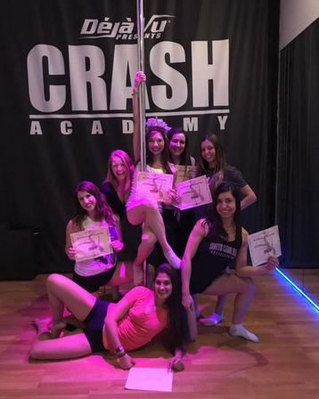 Group of women celebrating a pole fitness class graduation in a dance studio, posing around a vertical pole on a wooden floor under pink-purple lighting and holding certificates in front of a large backdrop.