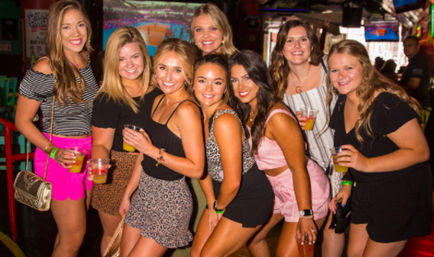Eight smiling young women in summer outfits posing with cocktails at a lively indoor bar — friends' night out, party vibes and nightlife.