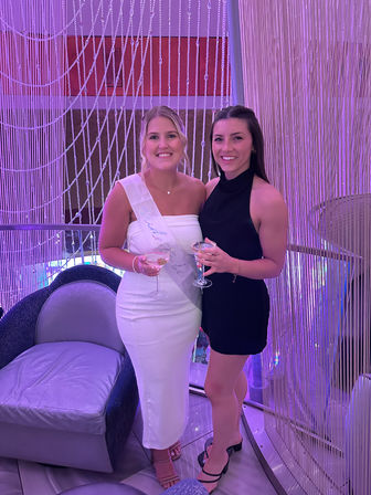 Two smiling women at a purple-lit indoor party, one wearing a white dress and sash and the other in a black dress, holding cocktails in front of a sparkling beaded curtain — bachelorette celebration vibe.
