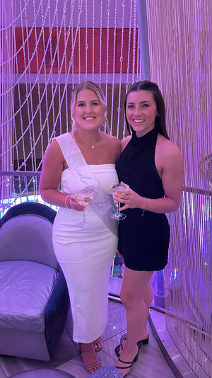 Two smiling women at a purple-lit indoor party, one wearing a white dress and sash and the other in a black dress, holding cocktails in front of a sparkling beaded curtain — bachelorette celebration vibe.