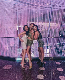 Three women in sequined cocktail dresses and heels smiling and posing together against a cascading beaded chandelier and pink-purple LED-lit backdrop in an indoor event space