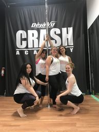 Five women in a pole-dance fitness class posing around a chrome pole in an indoor studio, wearing matching team tank tops on a hardwood floor.