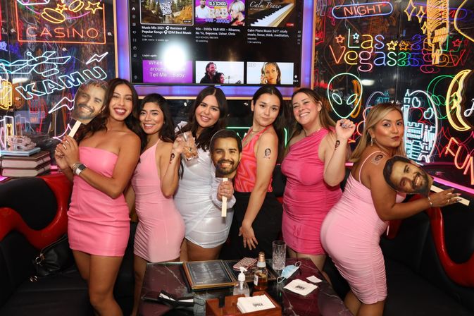 Group of seven women in pink and white dresses posing at a neon-lit Las Vegas nightclub, holding bearded-face photo props with drinks and menus on the table
