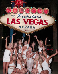 Cheerful group of women in white outfits posing and cheering beneath the illuminated Welcome to Fabulous Las Vegas, Nevada sign at night