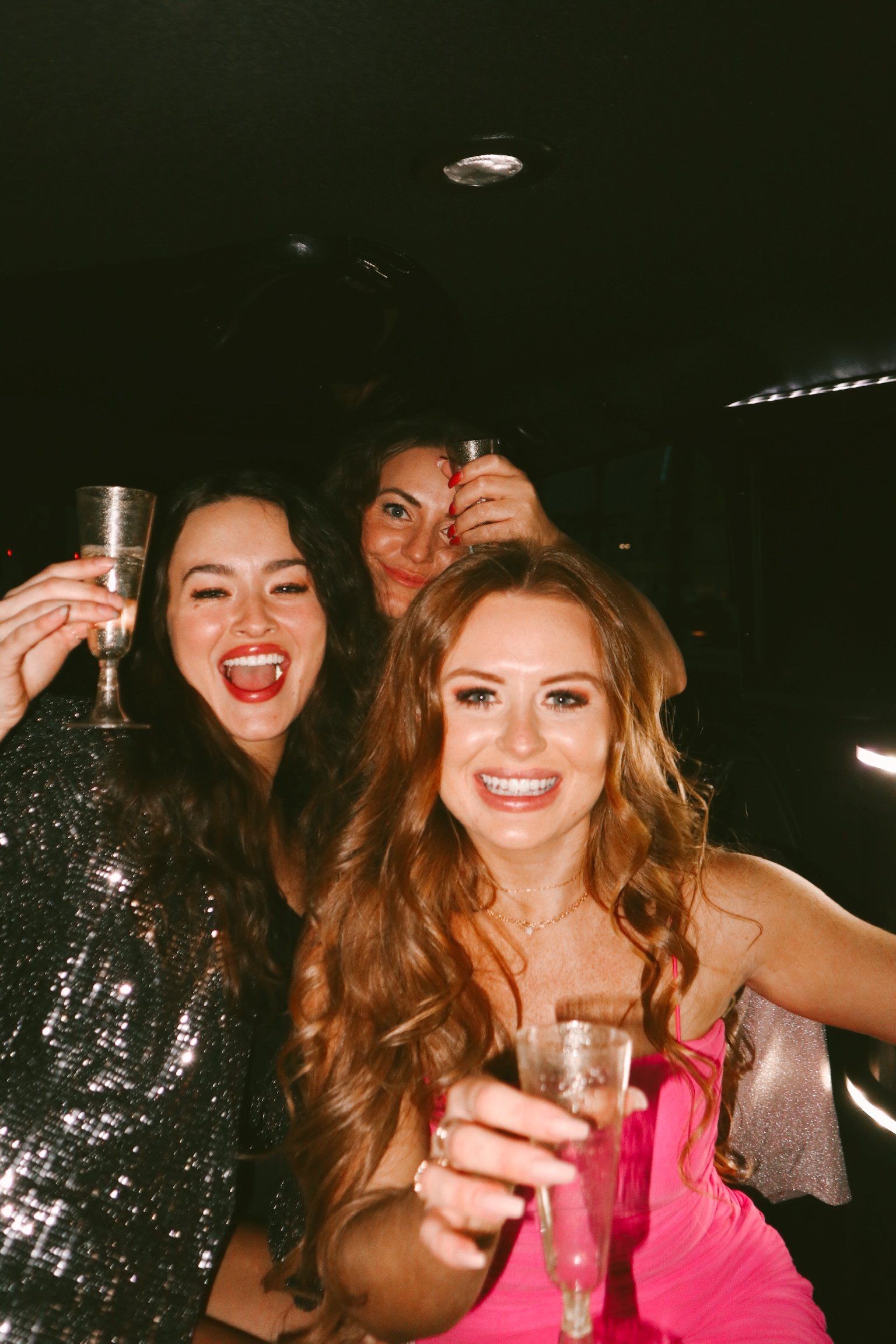 Three women smiling and toasting champagne in a dim limousine, one in a pink dress and another in sequins — lively girls’ night out and nightlife celebration.