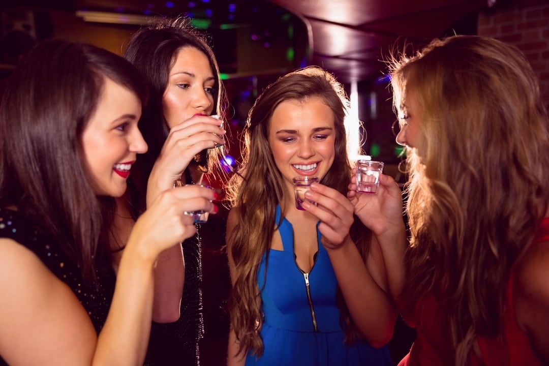 Four young women enjoying a girls' night out, smiling and raising shot glasses at a lively bar with colorful club lighting.