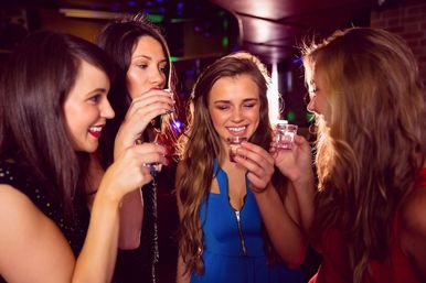 Four young women enjoying a girls' night out, smiling and raising shot glasses at a lively bar with colorful club lighting.