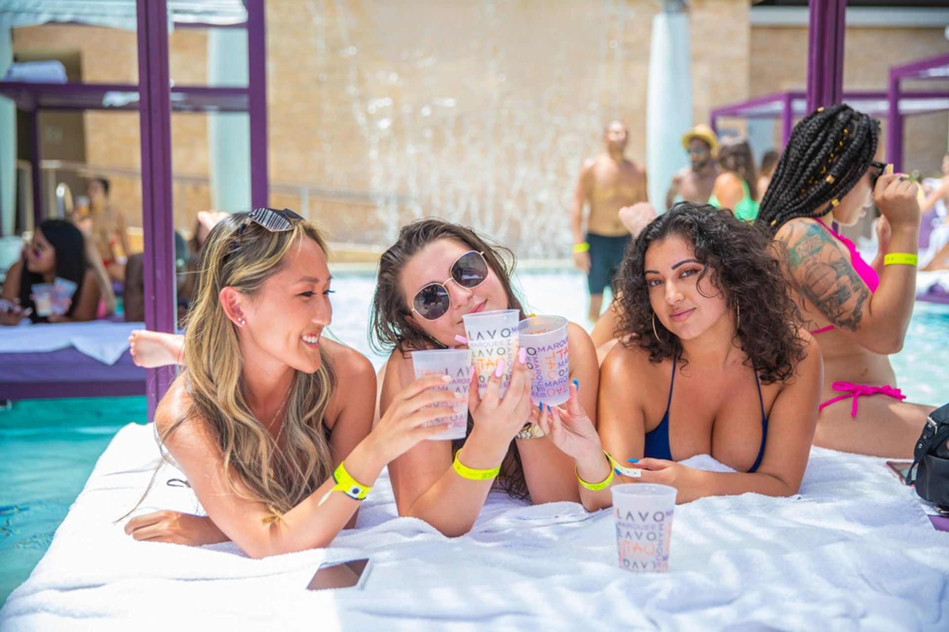 Three women smiling and clinking plastic cups while lounging on a towel in a sunny poolside cabana at a lively summer pool party with a waterfall feature in the background.