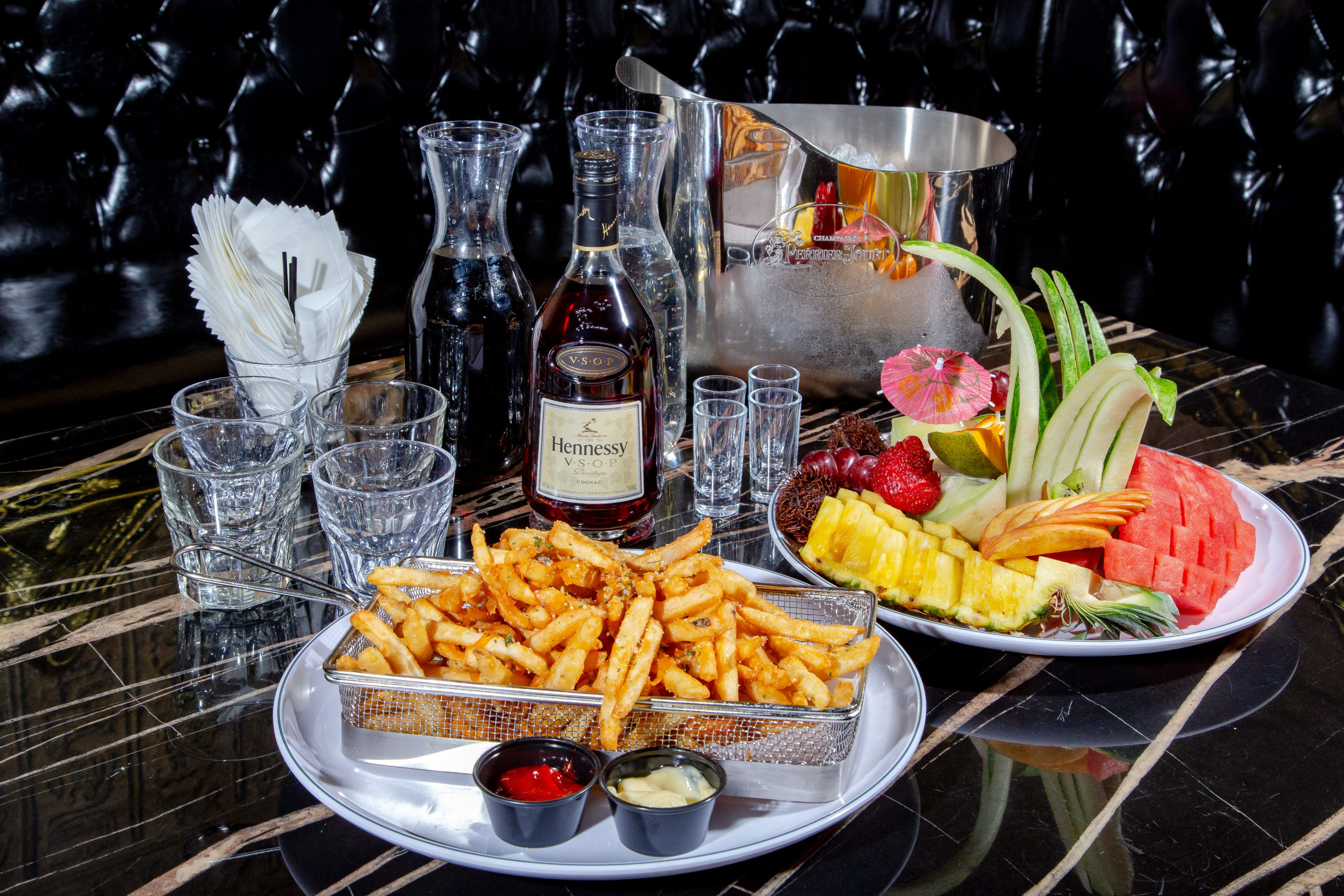 Seasoned fries in a metal basket with ketchup and mayo, colorful tropical fruit platter (pineapple, watermelon, melon, apple, strawberries), a bottle of cognac, shot glasses, ice bucket and carafes on a glossy marble lounge table.