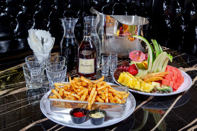 Seasoned fries in a metal basket with ketchup and mayo, colorful tropical fruit platter (pineapple, watermelon, melon, apple, strawberries), a bottle of cognac, shot glasses, ice bucket and carafes on a glossy marble lounge table.
