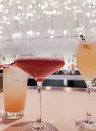 Three vibrant cocktails on a glossy bar counter—peach highball, deep red coupe, and light peach flute—against sparkling bokeh lights in a modern cocktail lounge