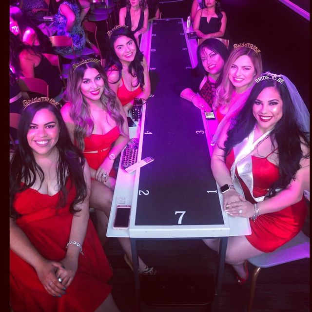 Smiling group of women in red dresses gathered around a numbered stage table under pink club lights at a bachelorette party, with the bride-to-be wearing a veil and sash.