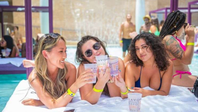 Three women in swimsuits lounging on a resort pool cabana, smiling and clinking plastic cups in a sunny daytime pool party with a fountain backdrop and wristbands.