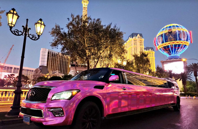 Pink stretch SUV limousine on the Las Vegas Strip at dusk, reflecting neon lights with ornate street lamps, palm trees, hotels and a colorful illuminated balloon-shaped sign in the background.
