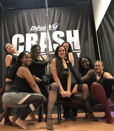 Seven women in a pole-dance fitness class pose together in black dancewear and stockings around a pole and chair inside a dance studio with a hardwood floor and dark backdrop.
