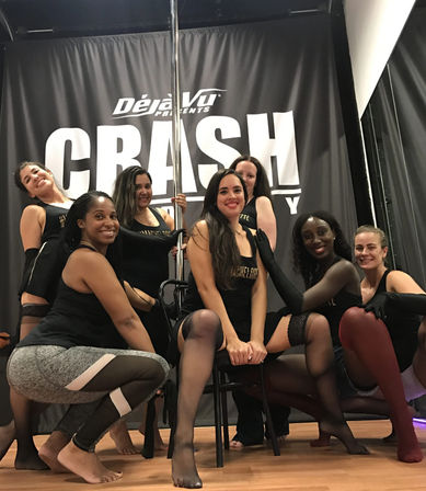 Seven women in a pole-dance fitness class pose together in black dancewear and stockings around a pole and chair inside a dance studio with a hardwood floor and dark backdrop.