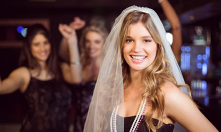 Smiling bride-to-be wearing a veil and pearl necklaces at a lively bachelorette party in a dimly lit bar with friends dancing in the background.